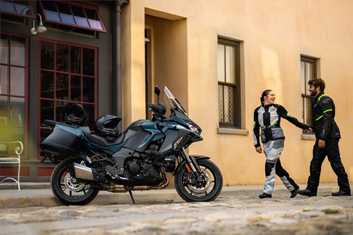Side angle of a parked motorcycle with people in the background walking. opens in a new window