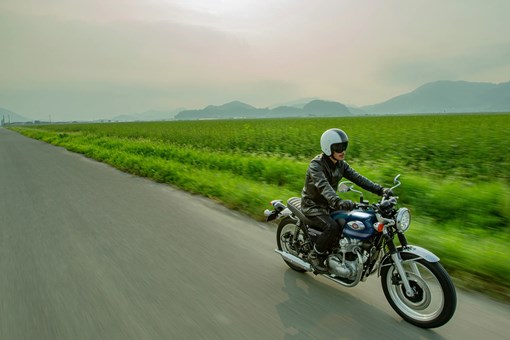 Three-quarter front angle of a person riding a motorcycle next to a field. opens in a new window