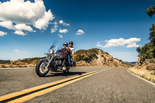 Three-quarter front angle of two people riding a motorcycle on a desert highway. opens in a new window