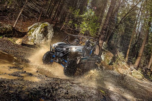 Three-quarter front angle of a person and passenger driving a side x side off-road through a puddle. opens in a new window