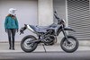 Side angle of a person standing next to a parked motorcycle on a city street.