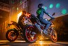 Three-quarter rear angle of two people seated on two separate motorcycles in a garage.