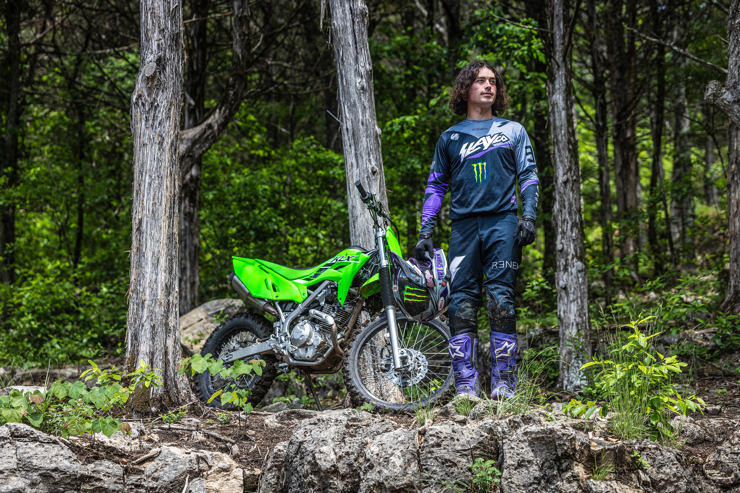 Front angle of a person standing next to a motorcycle with trees in the background.