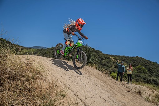 Side angle of a young adult riding an electric balance bike down a dirt hill.  opens in a new window