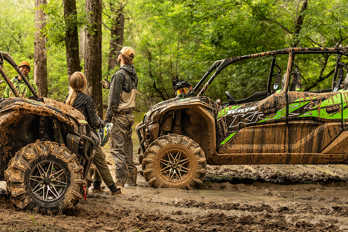A group of people talking between two side x sides parked off-road in the forest.