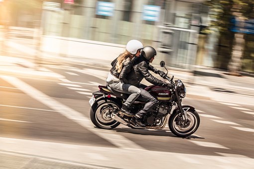 Side angle of a person and a passenger riding a motorcycle on a city street. opens in a new window