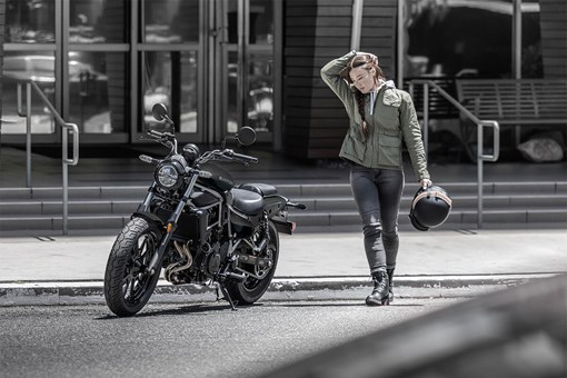  Three-quarter front angle of a person standing next to a motorcycle in front of a building. opens in a new window