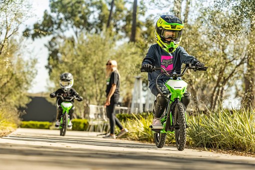 Three-quarter front angle of two kids riding electric balance bikes down a neighborhood street with a parent supervising. opens in a new window