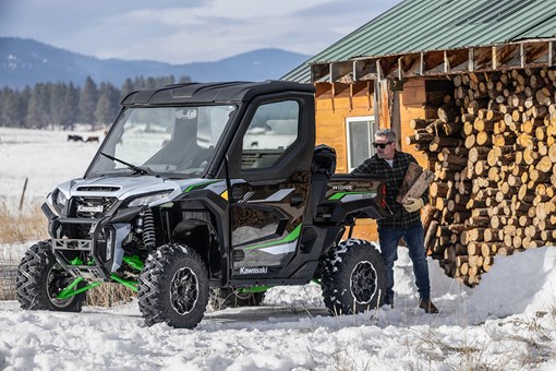 Front three-quarter view of a side x side parked in the snow next to a person. opens in a new window