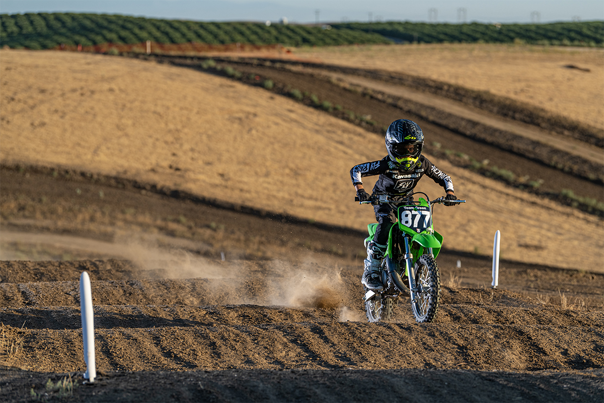 Three-quarter front angle of a person riding a green motorcycle on a dirt track.