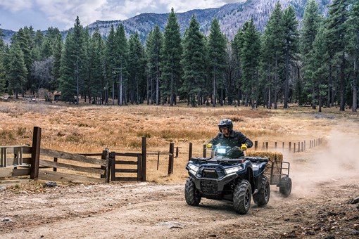 Three-quarter front angle of a person riding an ATV off-road on a large property with a trailer in tow. opens in a new window
