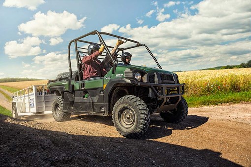 Three-quarter front angle of three people riding in a side x side on a ranch with a trailer in tow. opens in a new window