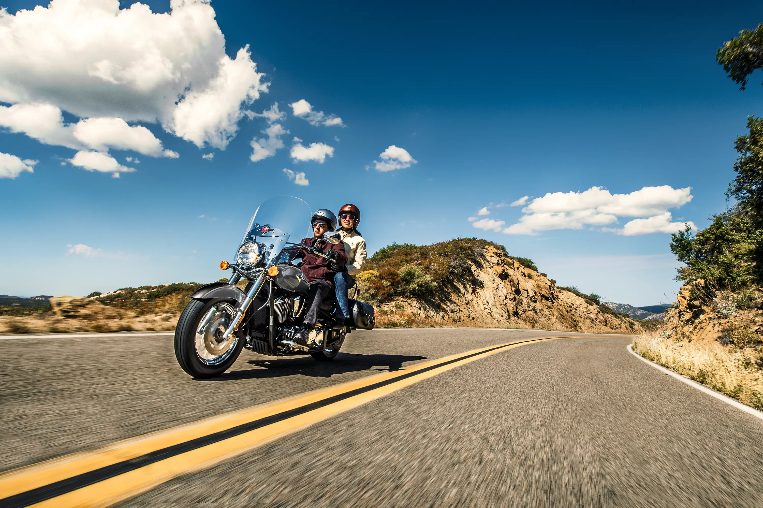 Three-quarter front angle of two people riding a motorcycle on a desert highway.