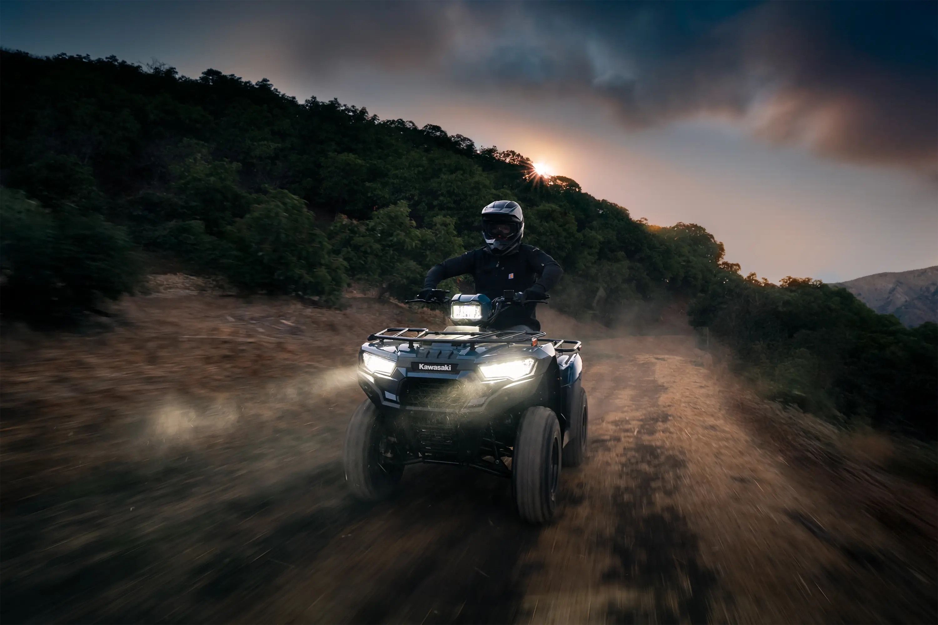 Front three-quarter angle of a person riding an ATV on a trail at sunset.
