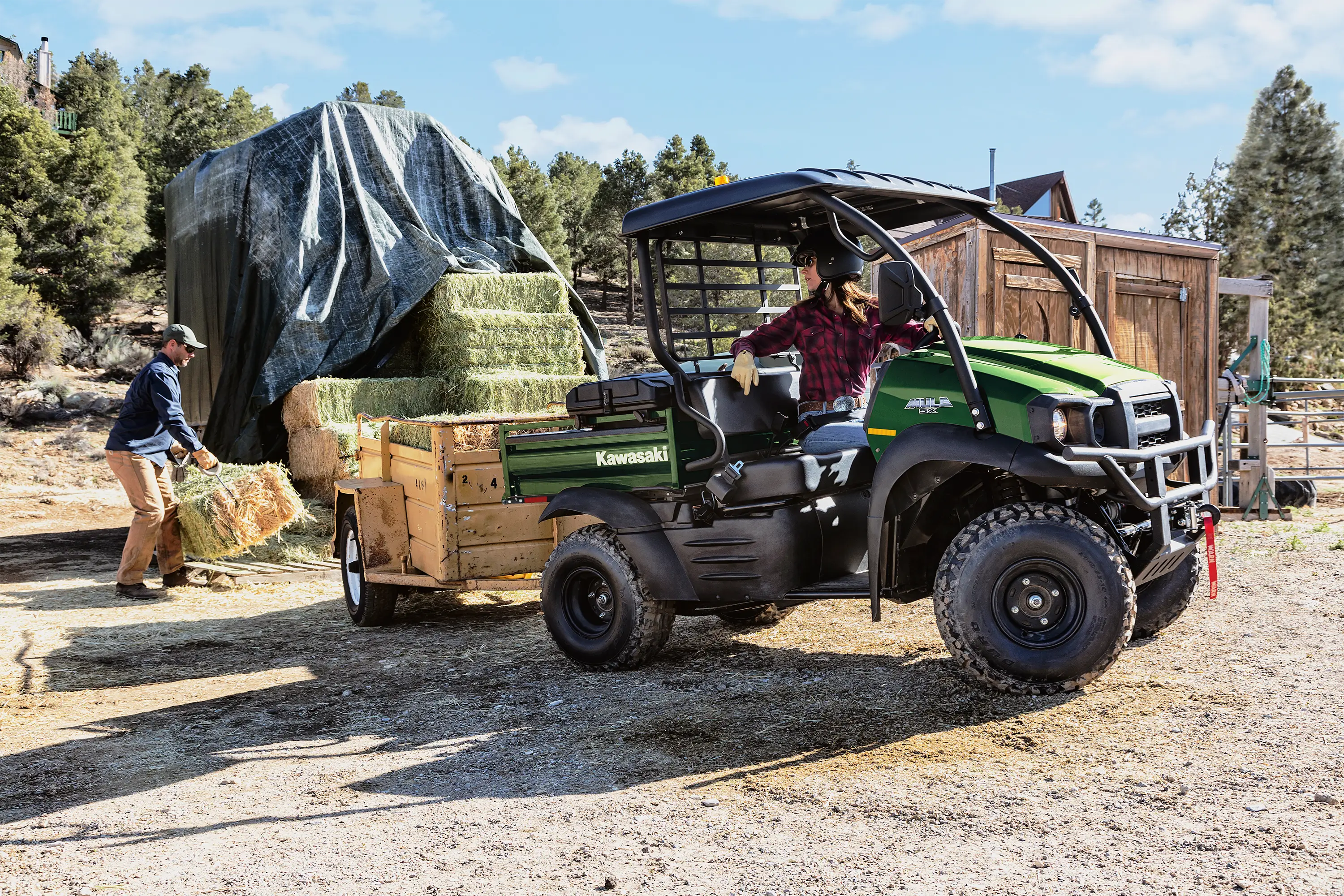 Side view of a person backing up a side x side with a trailer to a stack of hay bales.