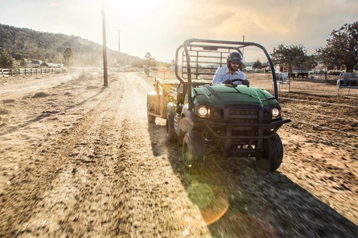 Front angle of a person driving a side x side through a field backlit by the sun. opens in a new window