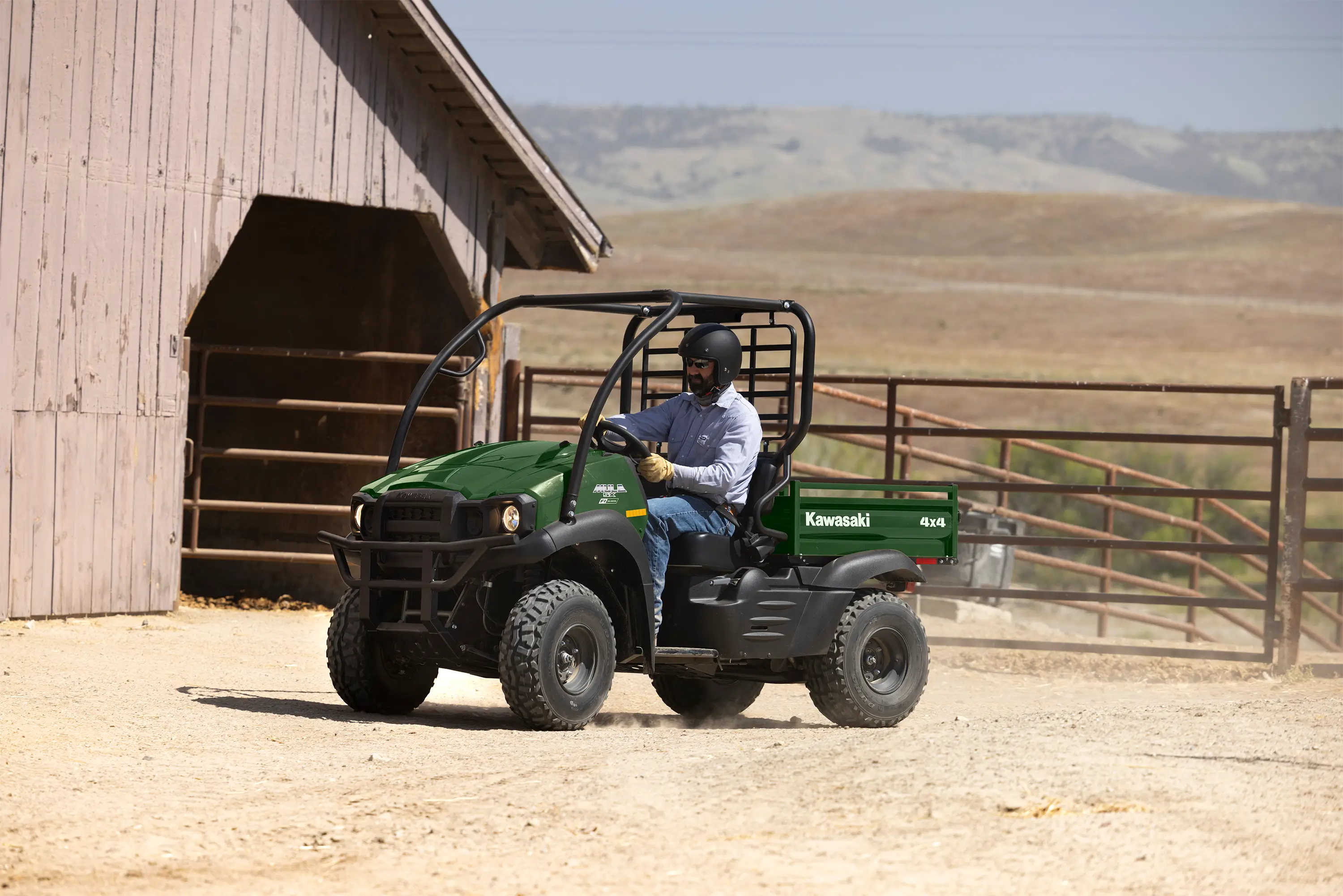 Front three-quarter view of a person driving a side x side near pipe corrals and a barn.