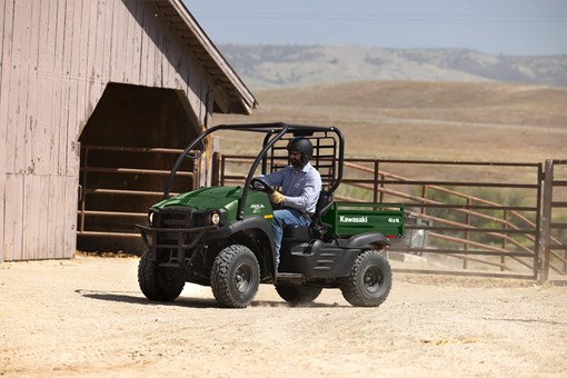 Front three-quarter view of a person driving a side x side near pipe corrals and a barn. opens in a new window