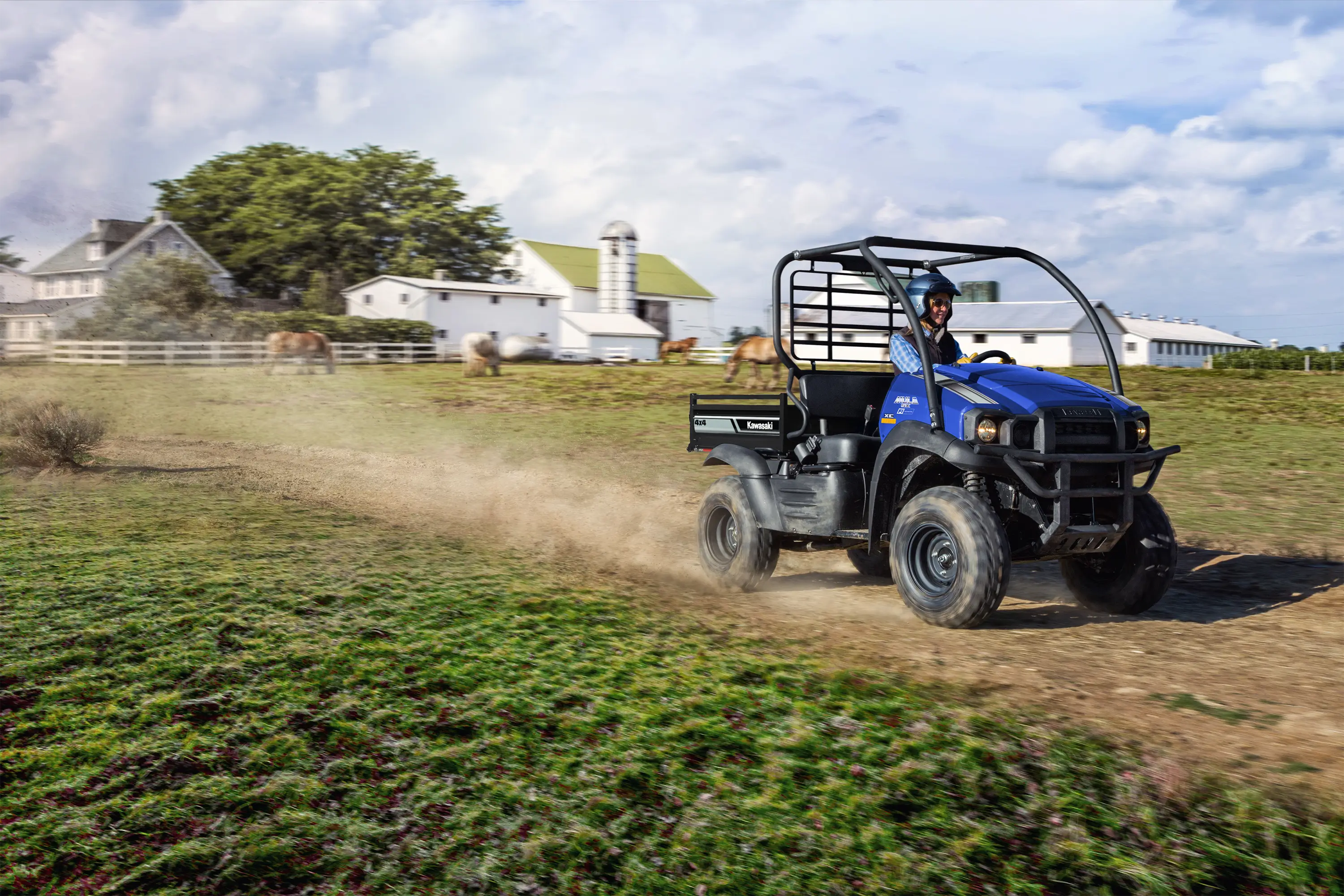Three-quarter view of a person driving a side x side on a dirt road with a white farmhouse in the background.