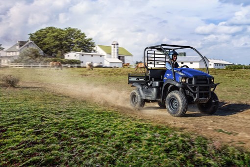 Three-quarter view of a person driving a side x side on a dirt road with a white farmhouse in the background. opens in a new window