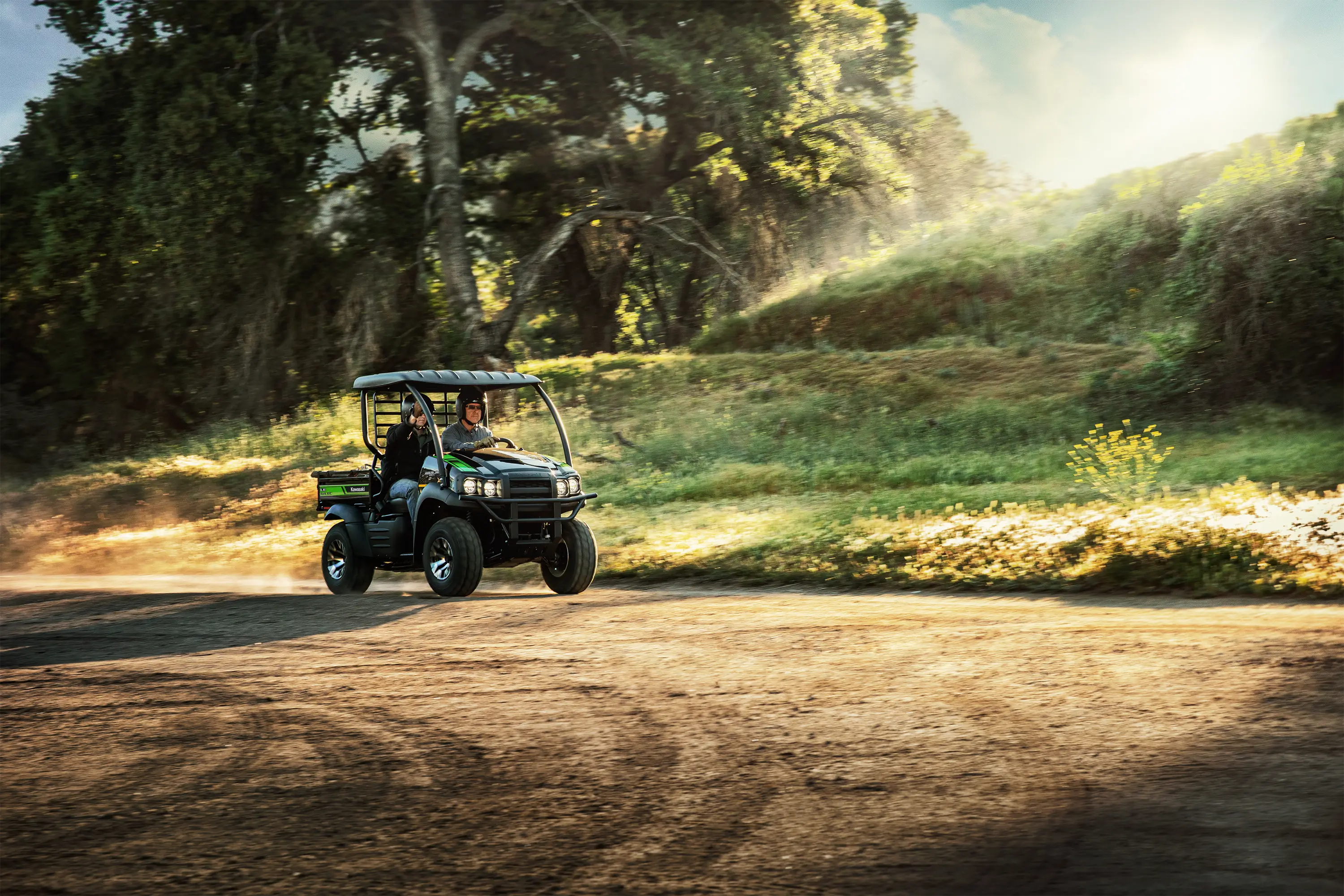 Two people riding in a side x side across a dirt lot with a tree in the background.