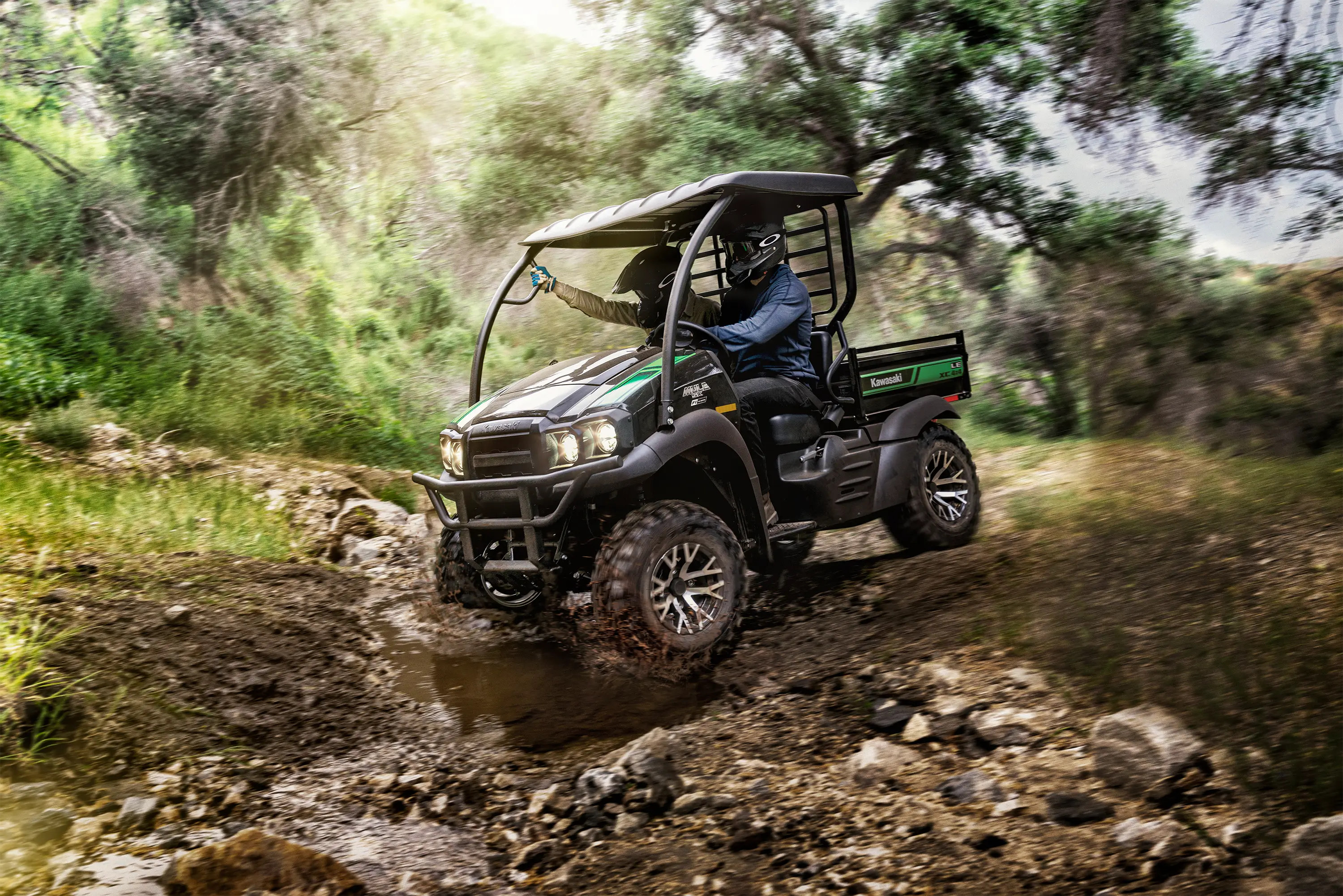 Action photo of two people riding in a side x side traversing a stream across a dirt road.