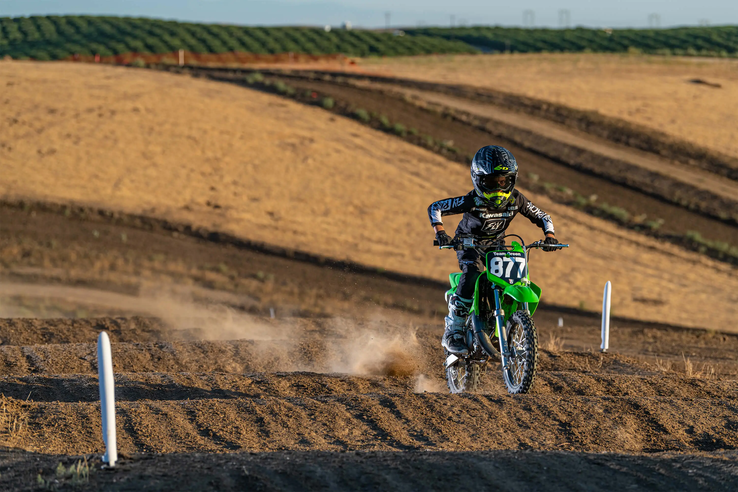 Front three-quarter right view of a kid riding a green youth dirt bike on a motocross track over bumps.