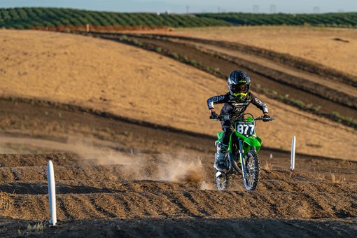 Front three-quarter right view of a kid riding a green youth dirt bike on a motocross track over bumps. opens in a new window