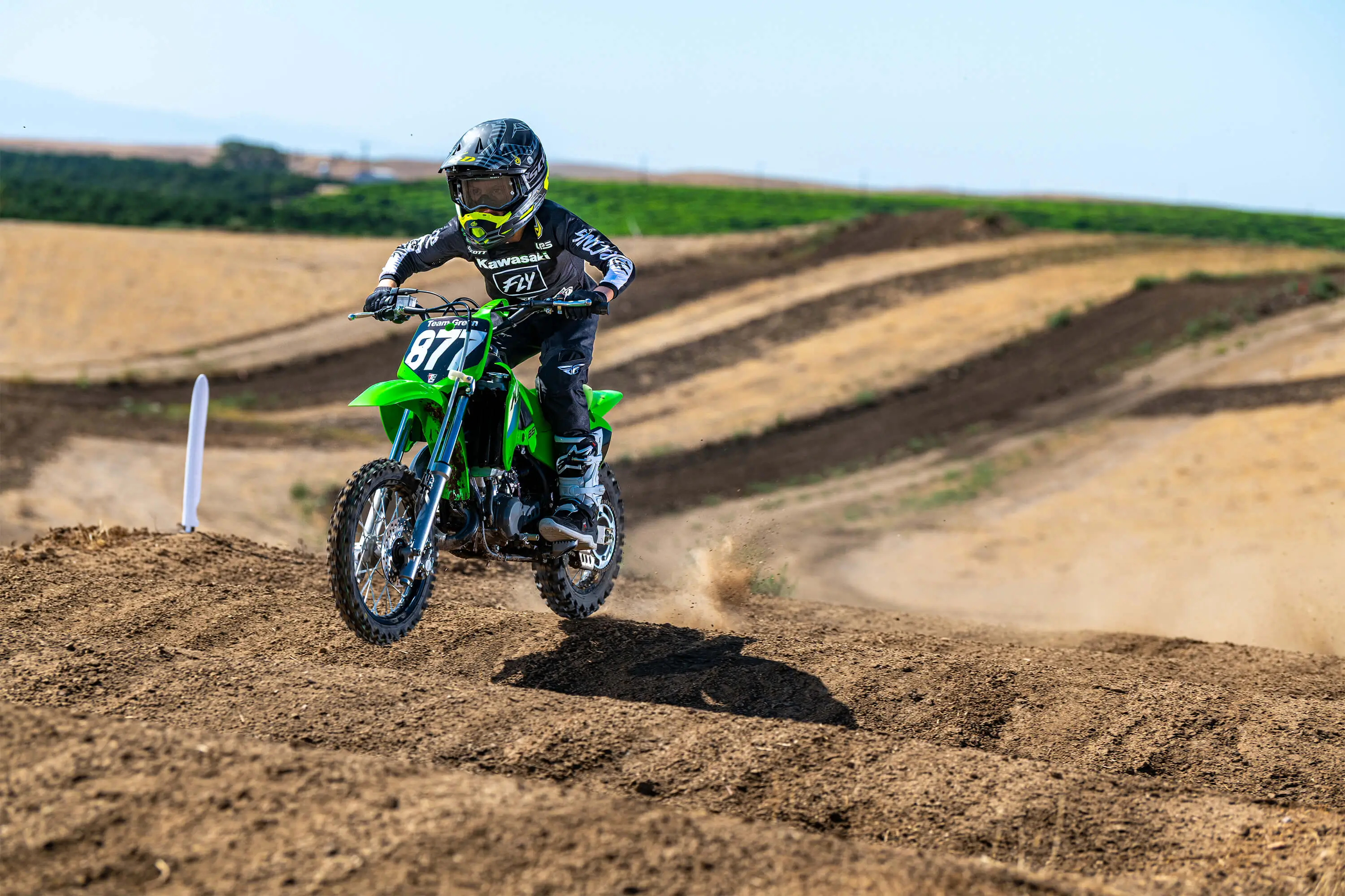 Front three-quarter left view of a kid riding a green youth motocross dirt bike on a track over bumps.