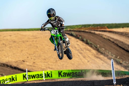 Oncoming view of a kid riding a green youth dirt bike over a jump on a motocross track. opens in a new window