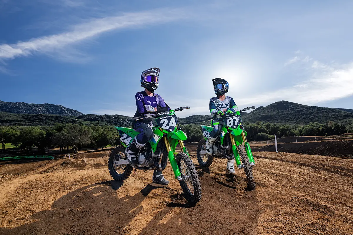 Two riders on green dirt bikes parked on a motocross track in front of a sunset.