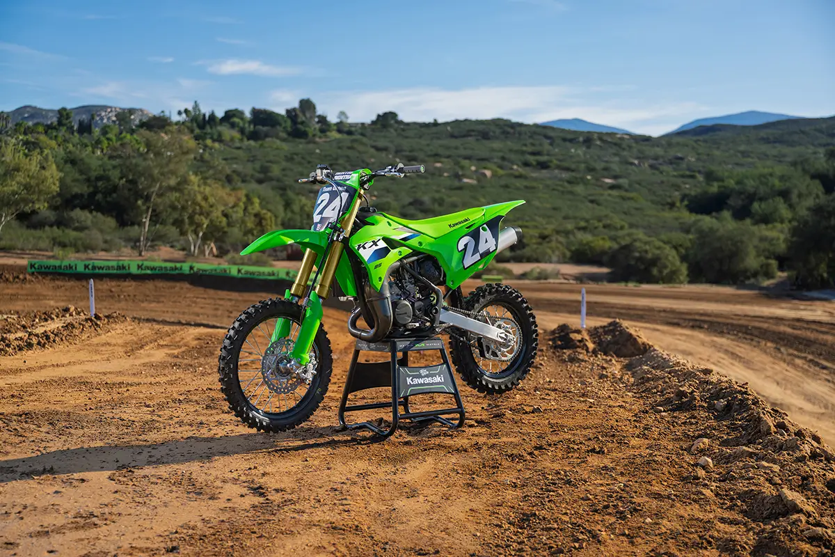 Front three-quarter view of a green youth dirt bike on a stand against a scenic outdoor background.