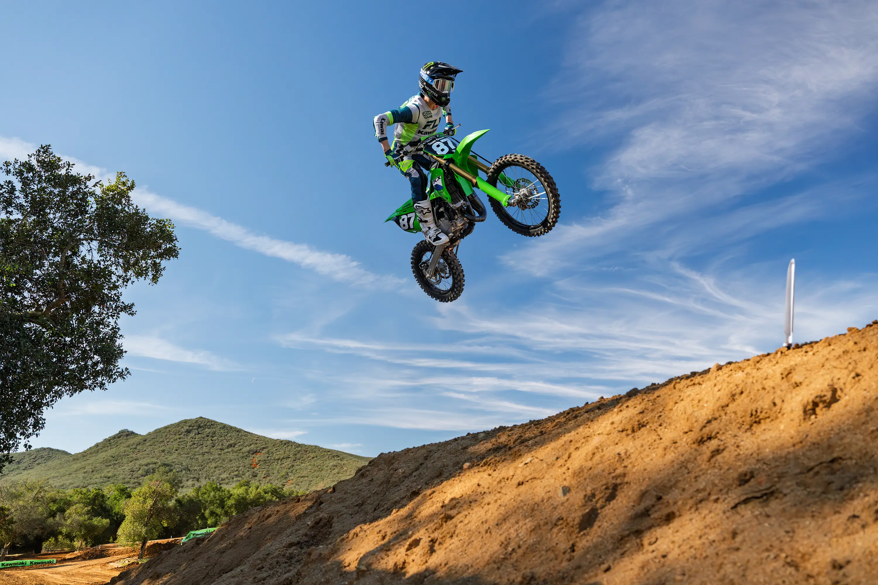 A person jumping a green dirt bike on a motocross track against a blue sky background.