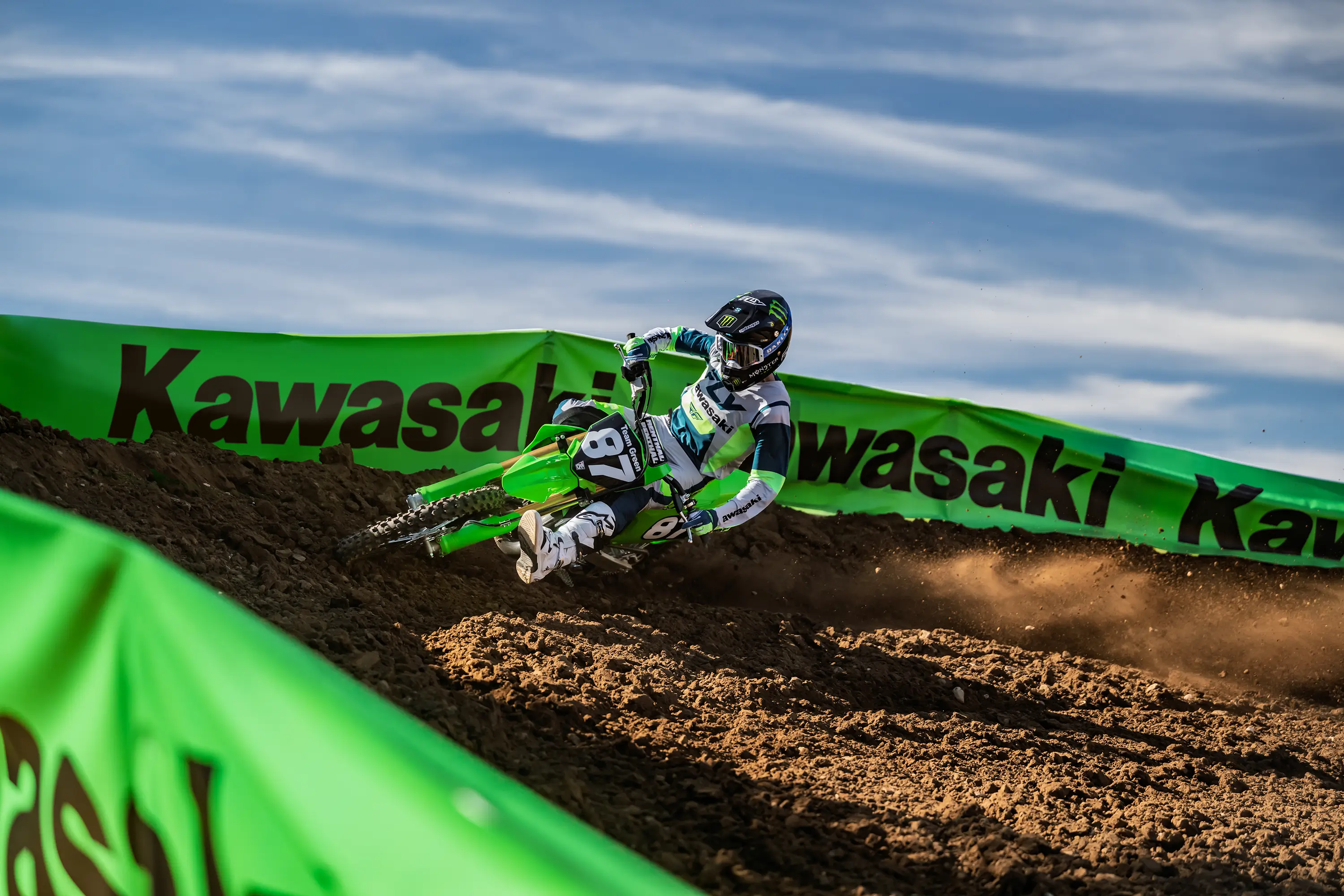 A person riding a green dirt bike fast around a turn with one foot out on a motocross track.