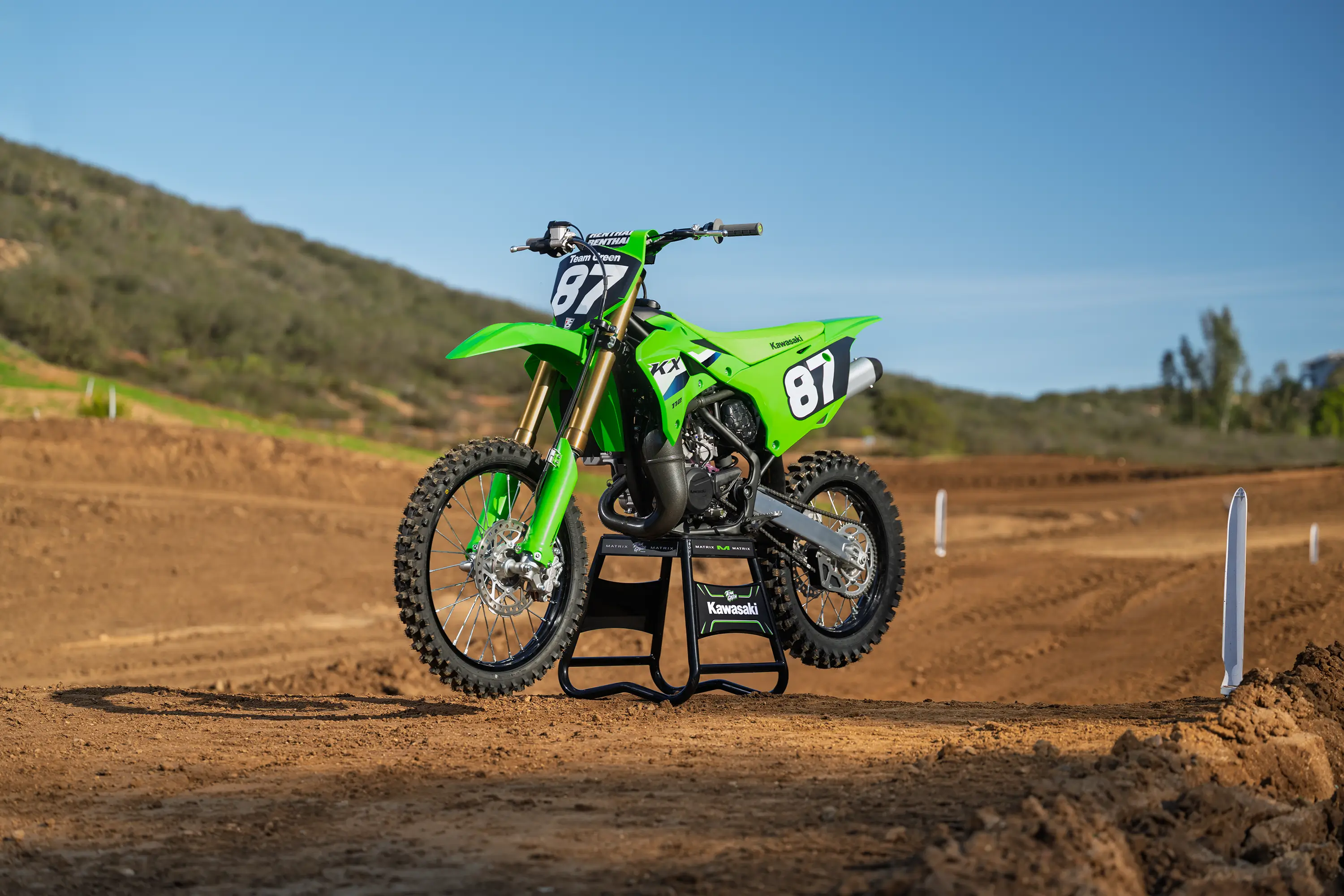 Front three-quarter view of a green dirt bike on a stand staged on a motocross track.