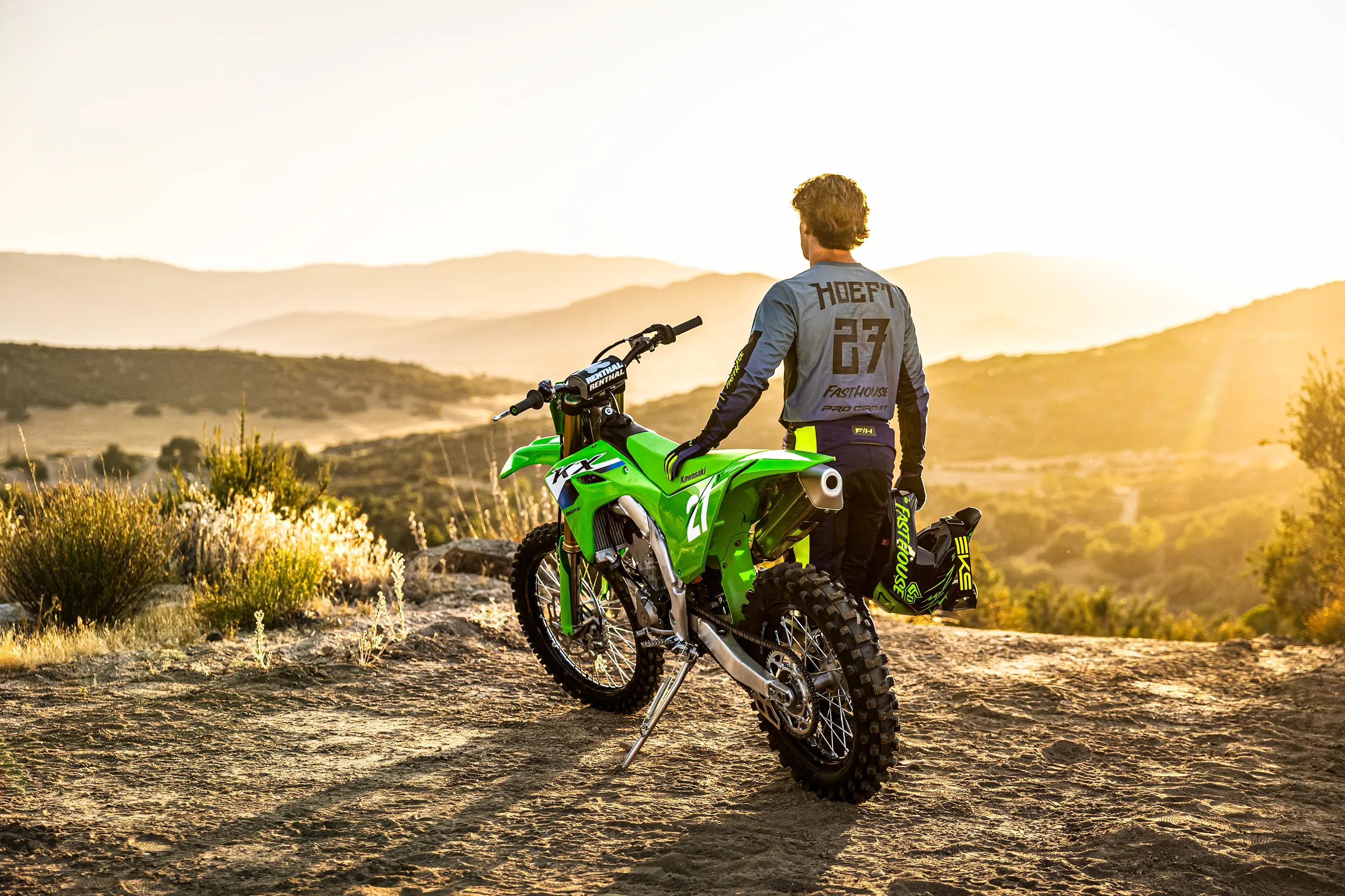 Three-quarter rear angle of a person standing next to a motorcycle parked at a dirt overlook.