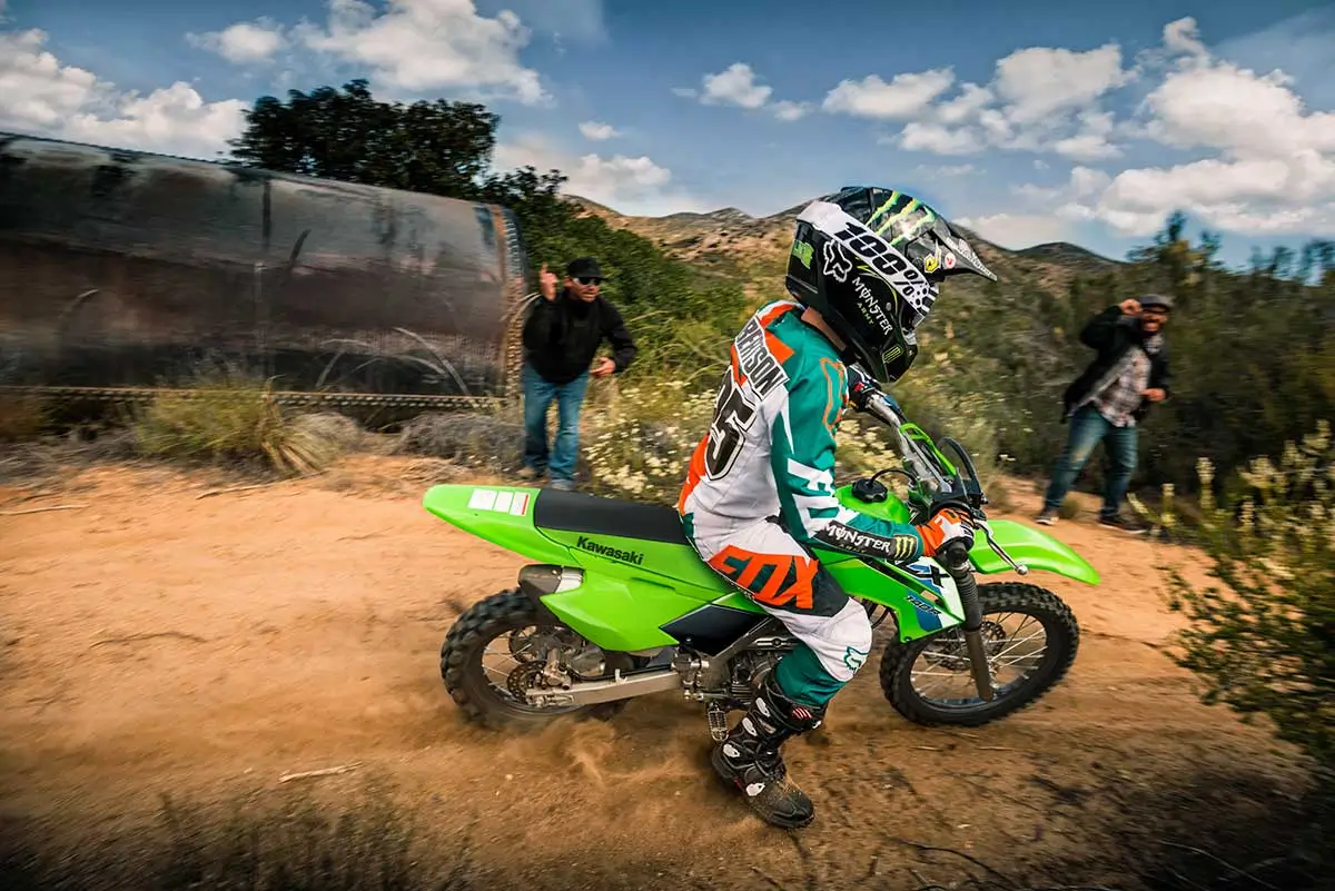 Side angle of a kid riding a dirt bike on a trail with a parent watching in the background.