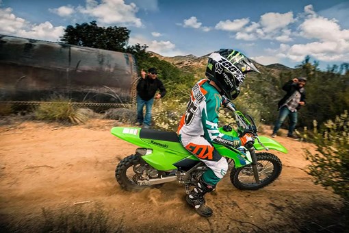Side angle of a kid riding a dirt bike on a trail with a parent watching in the background. opens in a new window