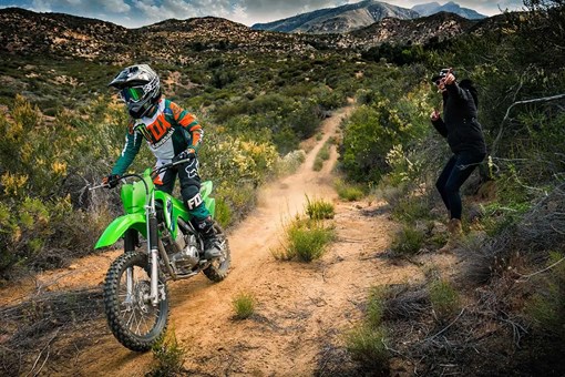 A kid riding a green dirt bike on a trail with a parent watching in the background. opens in a new window