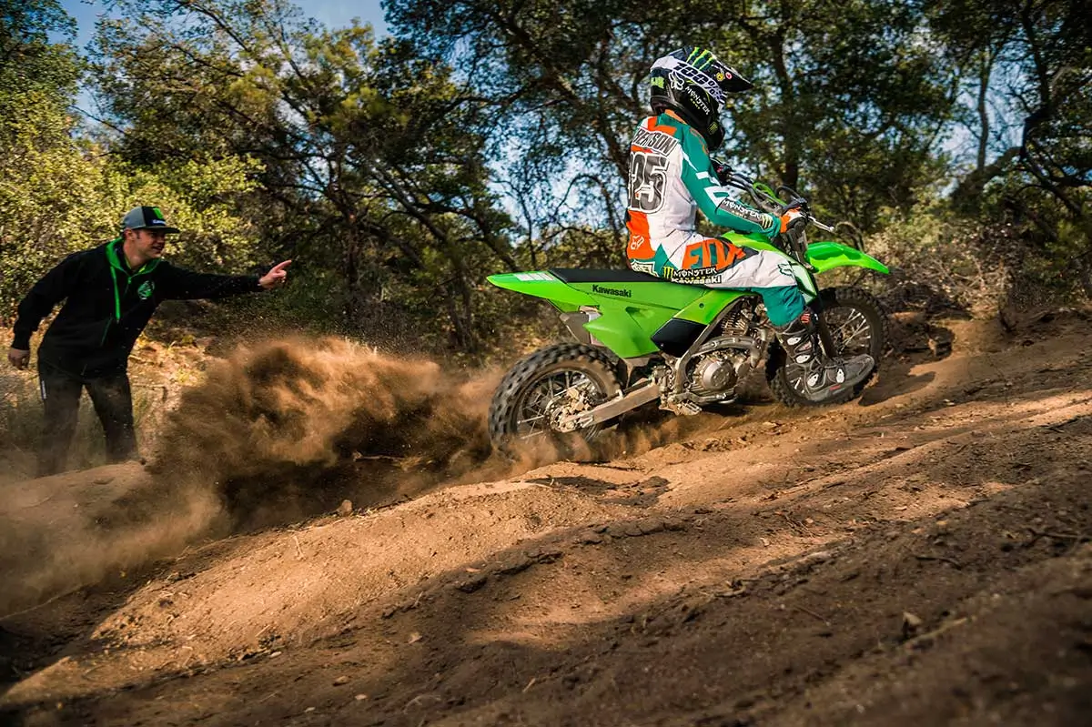 Side angle of a kid riding a dirt bike up a hill with a parent watching in the background.