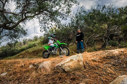 A kid a green dirt bike riding on a trail next to a parent. opens in a new window