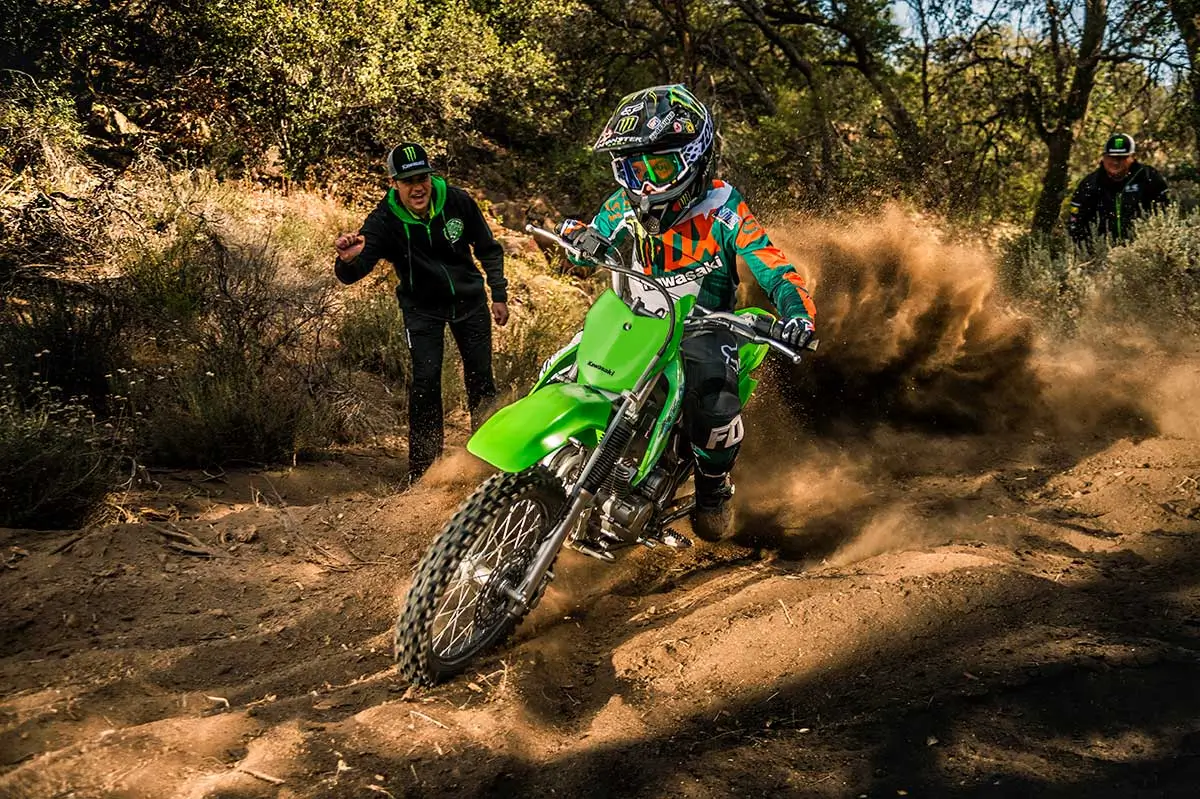 Oncoming view of a kid riding a dirt bike off-road with a parent watching in the background.