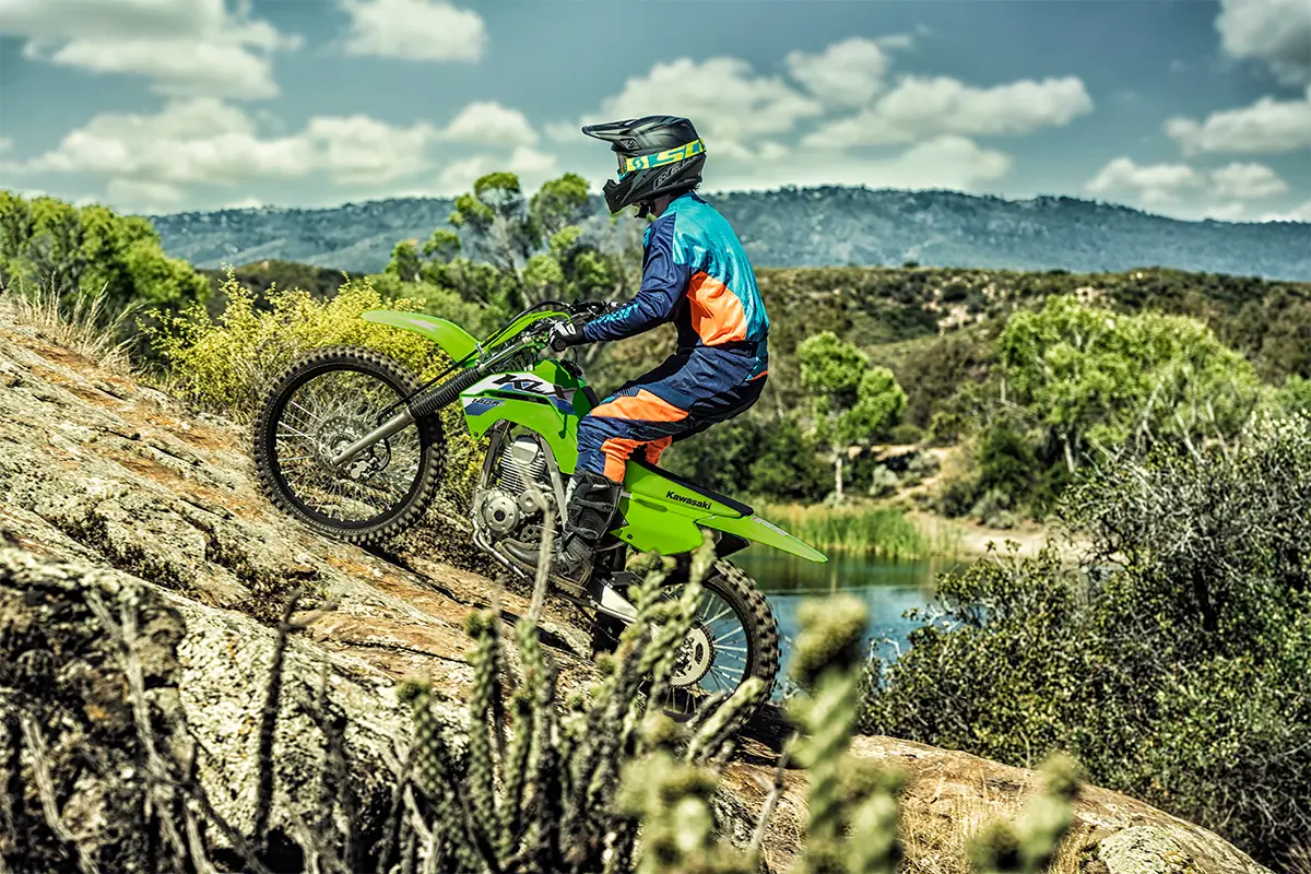 Side view of a person riding a green dirt bike up a hill in a forest.