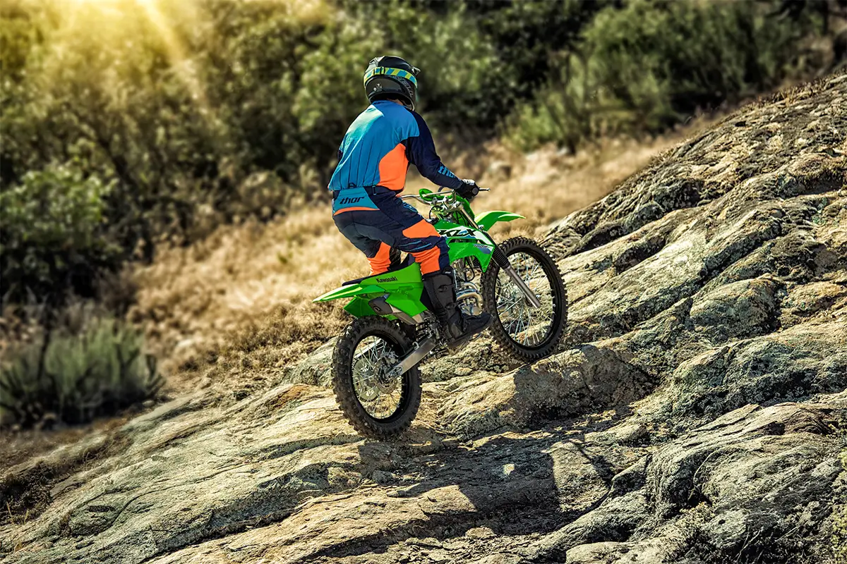 Rear three-quarter view of a person riding a green dirt bike up a rock face.