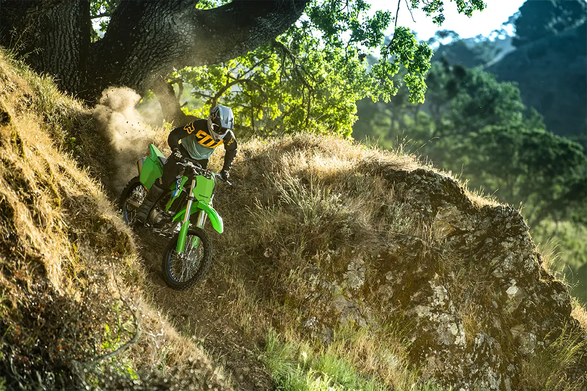 A person riding a green off-road dirt bike down a dusty hill under a tree.