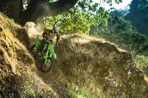 A person riding a green off-road dirt bike down a dusty hill under a tree. opens in a new window