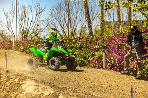 A kid riding a green youth ATV down a hill on a track as a parent gives encouragement. opens in a new window