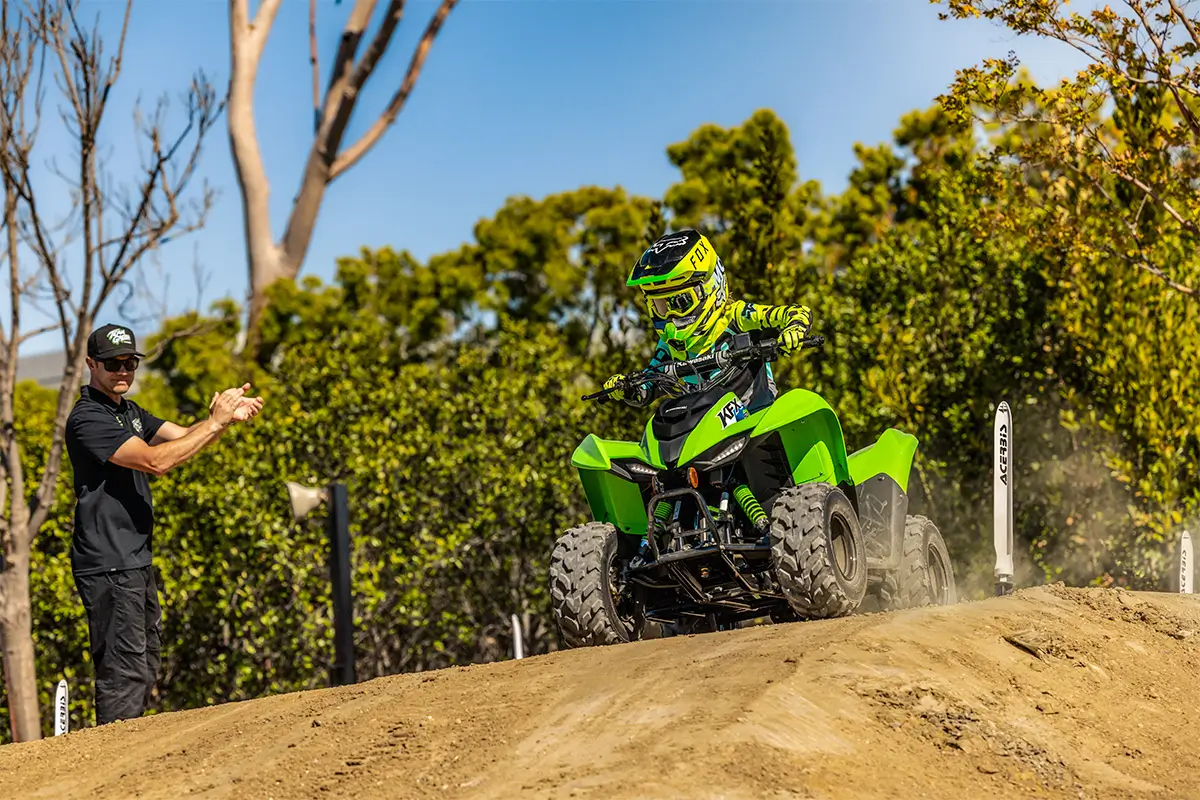 A kid riding a green youth ATV on a track as a parent gives instruction.