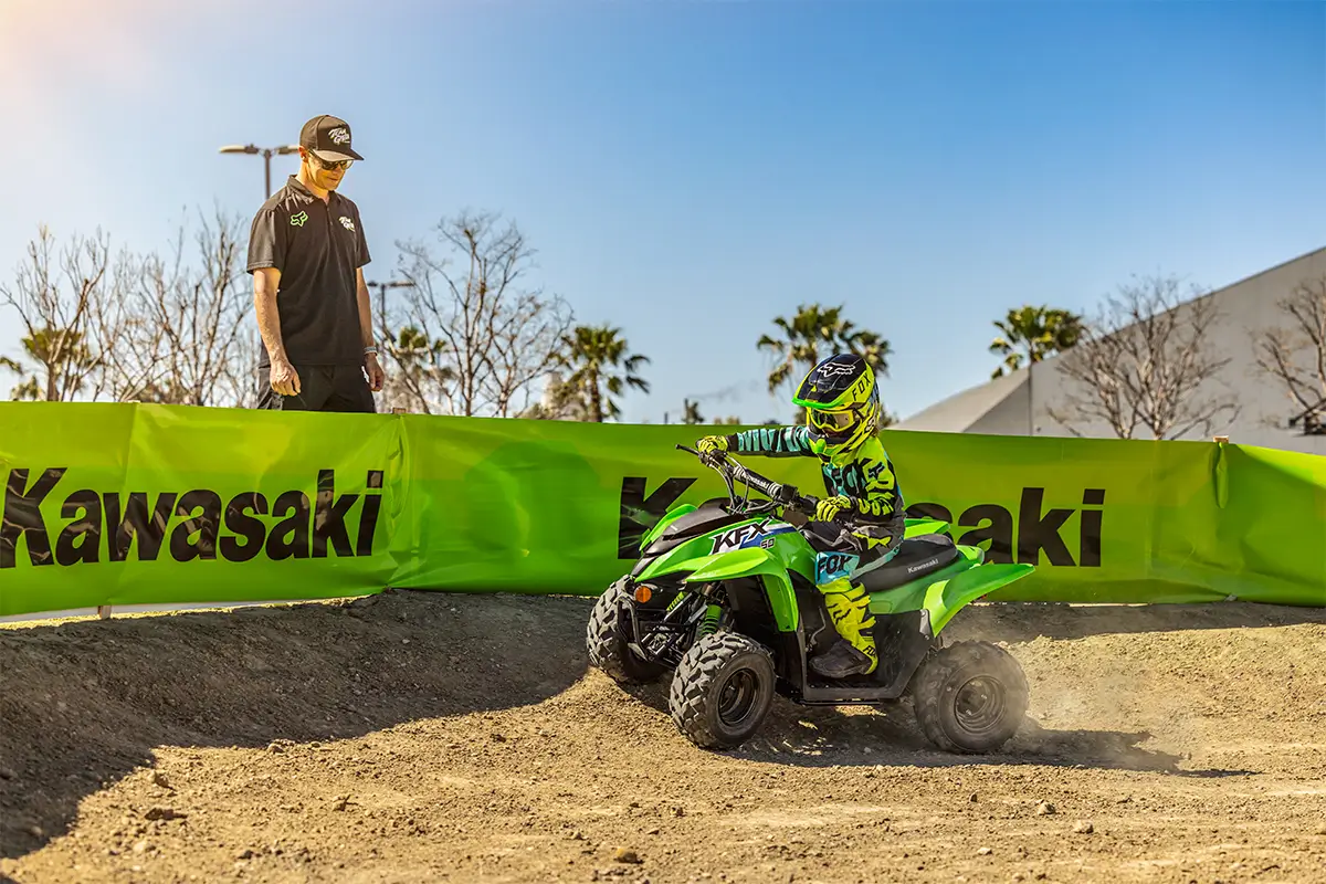 A kid riding a green youth ATV on a motocross track as a parent looks on.
