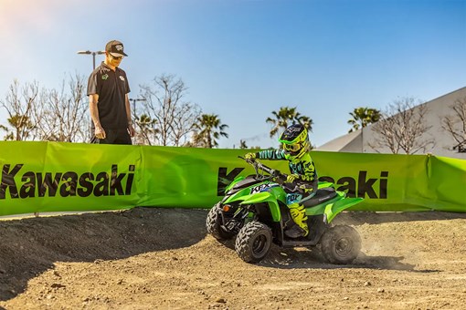 A kid riding a green youth ATV on a motocross track as a parent looks on. opens in a new window
